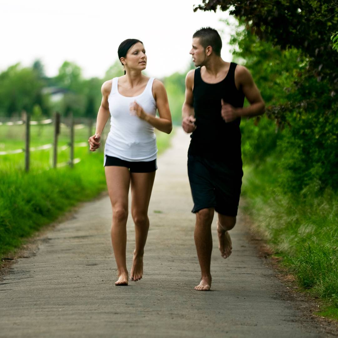 Ett par som springer barfota utomhus på en landsväg omgiven av grönska. Bilden speglar den naturliga rörelse, balans och frihetskänsla som NEO Sport barfotaskor från Nordic Rehab erbjuder — skor som stärker fotmuskulaturen, förbättrar gånghållning och stödjer en mer naturlig löpstil.