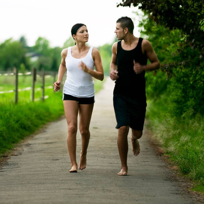Ett par som springer barfota utomhus på en landsväg omgiven av grönska. Bilden speglar den naturliga rörelse, balans och frihetskänsla som NEO Sport barfotaskor från Nordic Rehab erbjuder — skor som stärker fotmuskulaturen, förbättrar gånghållning och stödjer en mer naturlig löpstil.