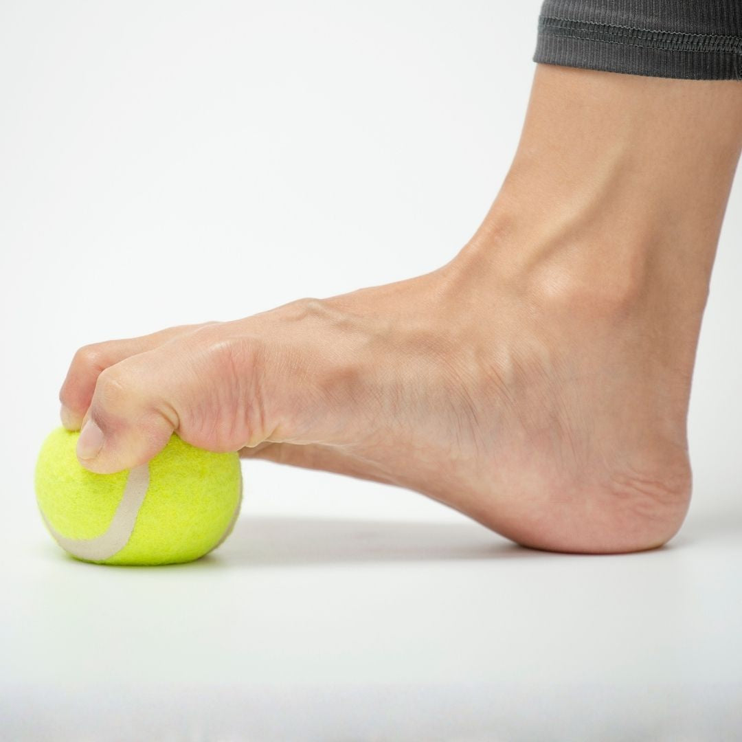 Närbild på en fot som greppar en tennisboll för att träna fotens muskler och rörlighet. Bilden symboliserar hur Nordic Rehab NEO Sport barfotaskor stärker fotmuskulaturen, förbättrar balans och ger naturlig stabilitet vid varje steg.